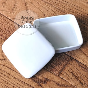 May include: White ceramic square box with a lid. The box is open and the lid is sitting next to it.
