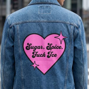 Puede incluir: Chaqueta vaquera azul claro con un gráfico de corazón rosa en la espalda. El corazón contiene el texto "Sugar, Spice, Fuck Ice" en letras negras, con dos estrellas. La chaqueta tiene un cuello clásico y cierre de botones.