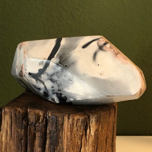 Può includere: Una scultura in ceramica bianca con segni astratti neri e grigi. La scultura ha la forma di una roccia geometrica e si trova su un ceppo di legno.
