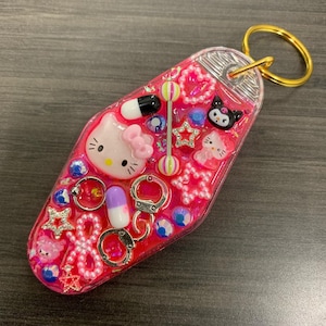 May include: Pink resin keychain with a Hello Kitty charm, a My Melody charm, a black and white Hello Kitty charm, and other charms including a pill, handcuffs, and a star. The keychain is shaped like a hotel key.