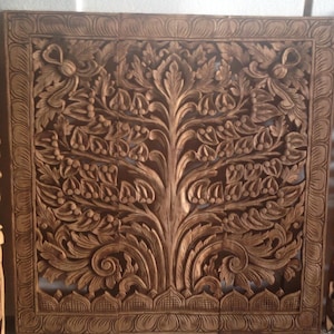 May include: A brown wooden wall hanging with a carved tree of life design. The tree is intricately detailed with leaves and branches, and the background is a solid brown.