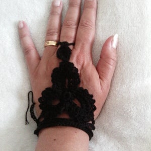 May include: A black crocheted fingerless glove with a delicate design. The glove is worn on a hand with a gold ring.