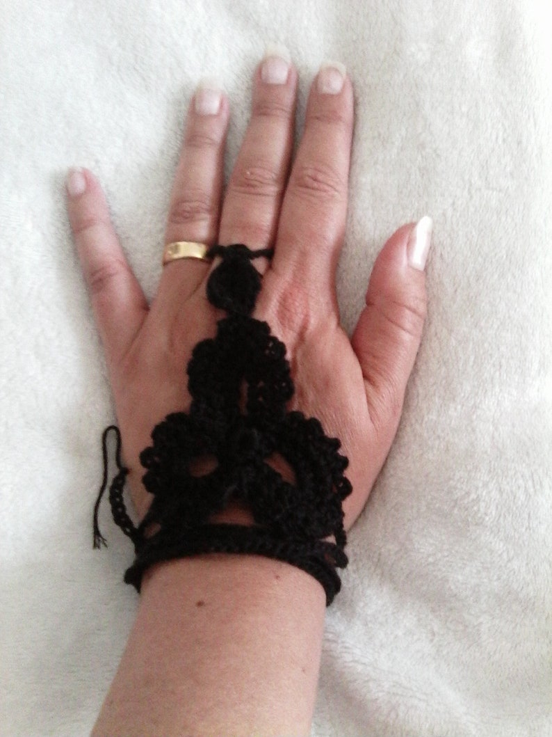 May include: A black crocheted fingerless glove with a delicate design. The glove is worn on a hand with a gold ring.