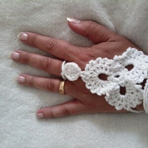 May include: White crocheted fingerless glove with a delicate floral design. The glove is worn on a hand with a gold ring.
