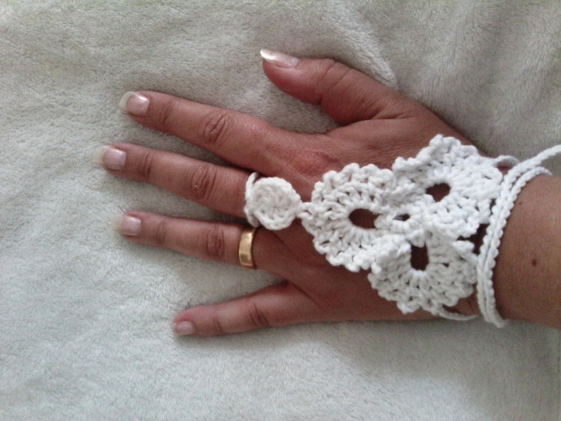 May include: White crocheted fingerless glove with a delicate floral design. The glove is worn on a hand with a gold ring.