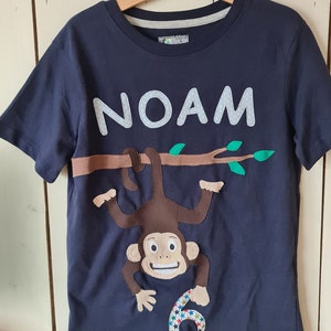May include: Navy blue t-shirt with the name "NOAM" in white block letters. A brown monkey hangs from a brown branch with green leaves. The number "6" is below the monkey, decorated with colorful stars.