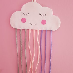 May include: A white cloud-shaped fabric with a smiling face, pink cheeks, and closed eyes. It has multiple fabric loops hanging from the bottom, in shades of gray, pink, and purple.