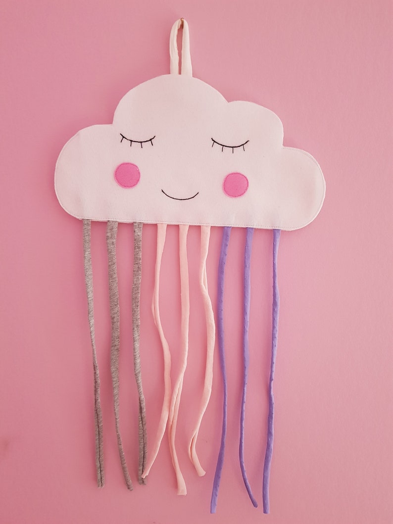 May include: A white cloud-shaped fabric with a smiling face, pink cheeks, and closed eyes. It has multiple fabric loops hanging from the bottom, in shades of gray, pink, and purple.