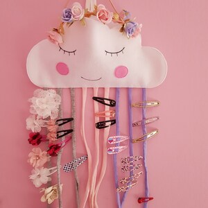 May include: A white felt cloud with a smiling face and a flower crown hanging on a pink wall. The cloud has multiple colorful hair clips hanging from it.