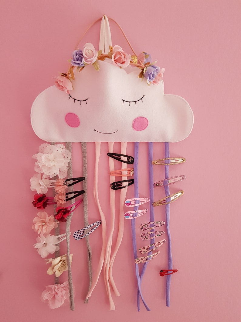 May include: A white felt cloud with a smiling face and a flower crown hanging on a pink wall. The cloud has multiple colorful hair clips hanging from it.
