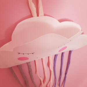 May include: A white felt cloud with a smiling face and closed eyes hangs from a pink wall.  The cloud has a pocket and is attached to colorful strings that resemble rain.