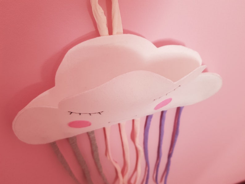 May include: A white felt cloud with a smiling face and closed eyes hangs from a pink wall.  The cloud has a pocket and is attached to colorful strings that resemble rain.