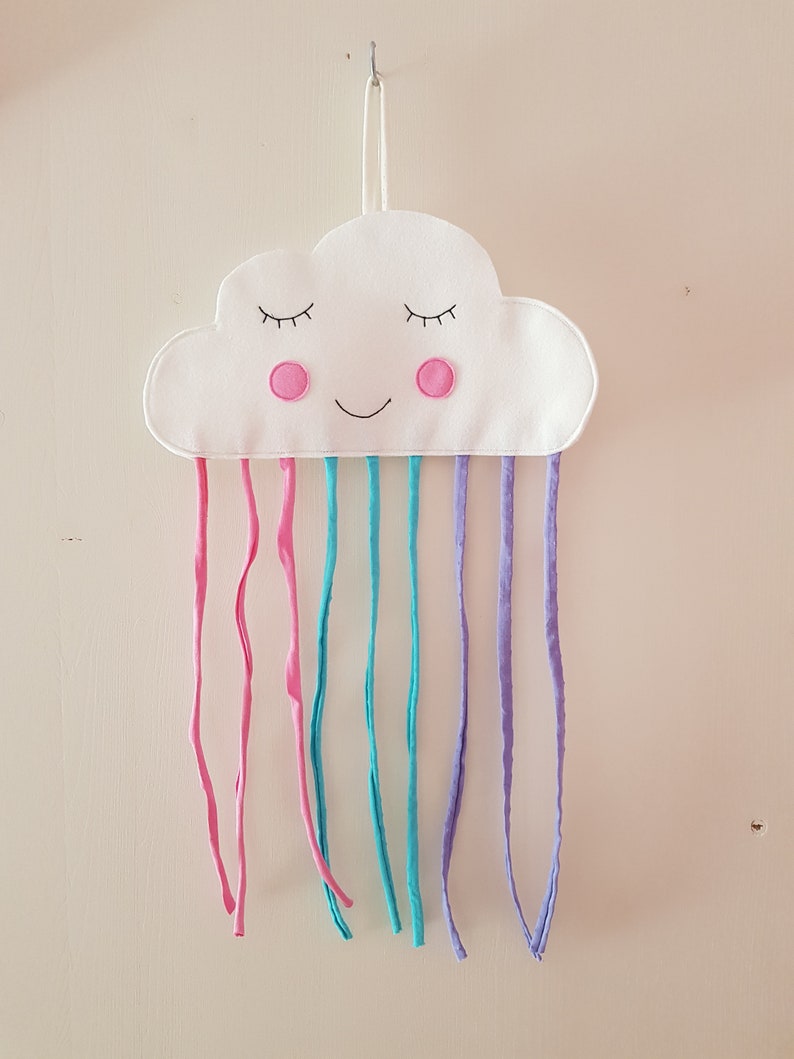 May include: A white felt cloud with a smiling face and pink cheeks, hanging from a hook.  It has rainbow-colored fabric strips hanging from the bottom.