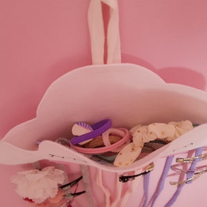 May include: A white felt cloud-shaped hair accessory organizer with a pink interior. The organizer is filled with various hair ties, clips, and scrunchies in pink, purple, and gold.