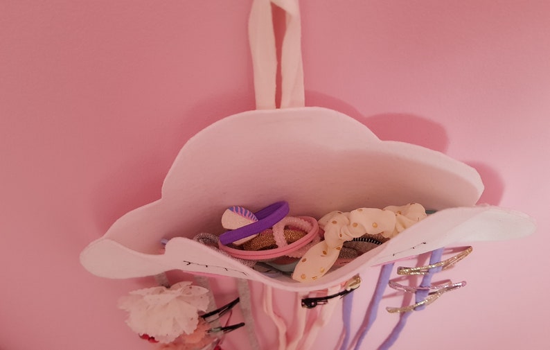 May include: A white felt cloud-shaped hair accessory organizer with a pink interior. The organizer is filled with various hair ties, clips, and scrunchies in pink, purple, and gold.