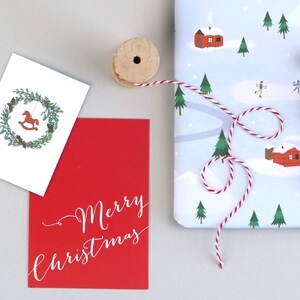 Puede incluir: Una tarjeta roja con texto blanco en cursiva que dice "Merry Christmas". Una tarjeta blanca con una corona verde y un caballo de balanc&iacute;n rojo est&aacute; en el fondo. Un carrete de hilo rojo y blanco est&aacute; sobre la mesa.