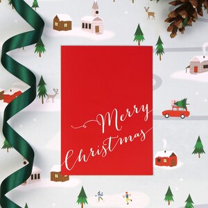 Puede incluir: Una tarjeta roja con texto blanco que dice "Merry Christmas" sobre un fondo blanco con una escena de invierno. La escena incluye casas, &aacute;rboles y un coche con un &aacute;rbol de Navidad en la parte superior.