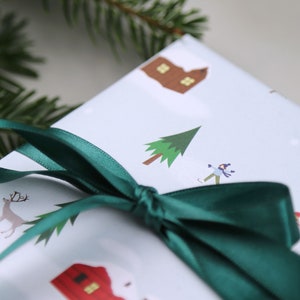 Puede incluir: Una caja de regalo blanca con una cinta verde atada alrededor. La caja est&aacute; decorada con una escena de invierno, que incluye una peque&ntilde;a casa, un &aacute;rbol, una persona patinando sobre hielo y un reno.