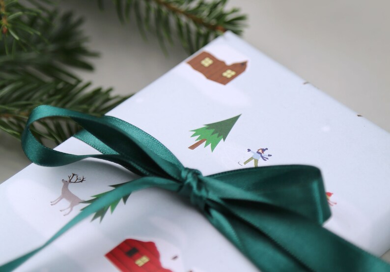 Puede incluir: Una caja de regalo blanca con una cinta verde atada alrededor. La caja est&aacute; decorada con una escena de invierno, que incluye una peque&ntilde;a casa, un &aacute;rbol, una persona patinando sobre hielo y un reno.