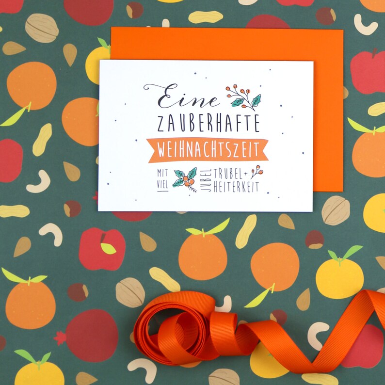 May include: A white card with the text "Eine zauberhafte Weihnachtszeit mit viel Jubel, Trubel & Heiterkeit" on a green background with orange and red illustrations of fruit and nuts. The card is tied with an orange ribbon.