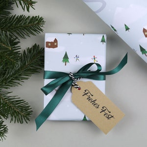 Puede incluir: Una caja de regalo blanca con una cinta verde y una etiqueta de regalo marr&oacute;n que dice "Frohes Fest". La caja est&aacute; envuelta en papel con una escena de invierno de casas, &aacute;rboles y personas esquiando.