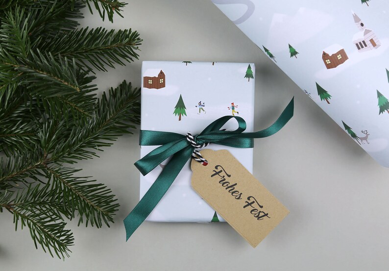 Puede incluir: Una caja de regalo blanca con una cinta verde y una etiqueta de regalo marr&oacute;n que dice "Frohes Fest". La caja est&aacute; envuelta en papel con una escena de invierno de casas, &aacute;rboles y personas esquiando.