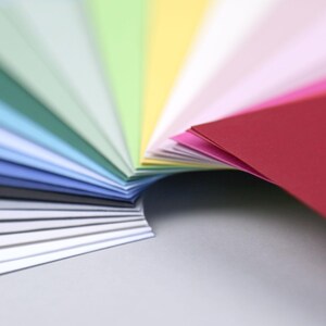 Op de afbeelding: Een waaier van kleurrijke papieren vellen, gerangschikt in een regenboogpatroon. De vellen zijn dubbelgevouwen en waaiervormig uitgespreid, waardoor een gelaagd effect ontstaat. De kleuren vari&euml;ren van donkerblauw tot rood.