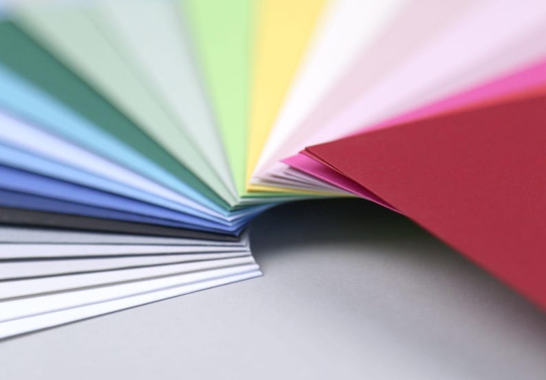 Op de afbeelding: Een waaier van kleurrijke papieren vellen, gerangschikt in een regenboogpatroon. De vellen zijn dubbelgevouwen en waaiervormig uitgespreid, waardoor een gelaagd effect ontstaat. De kleuren vari&euml;ren van donkerblauw tot rood.
