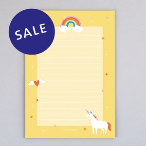 Könnte beinhalten: Gelber, linierter Notizblock mit einem Regenbogen, Wolken, Sternen, einem Herz mit Flügeln und einem Einhorn. Der Notizblock trägt die Aufschrift "SALE" in einem blauen Kreis.