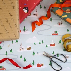 Puede incluir: Papel de regalo azul y blanco con una escena de pueblo de invierno. El papel presenta casas, &aacute;rboles, un coche con un &aacute;rbol de Navidad en la parte superior y un ping&uuml;ino. Hay una cinta roja, una cinta dorada y un par de tijeras negras en el papel.