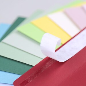 Op de afbeelding: Een rode envelop met een witte plakstrip, die gedeeltelijk is losgetrokken, waardoor de kleverige kant zichtbaar wordt. De envelop ligt op een stapel gekleurde vellen papier in tinten blauw, groen, geel en roze.