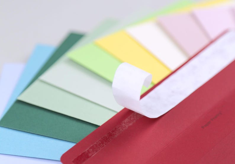 Op de afbeelding: Een rode envelop met een witte plakstrip, die gedeeltelijk is losgetrokken, waardoor de kleverige kant zichtbaar wordt. De envelop ligt op een stapel gekleurde vellen papier in tinten blauw, groen, geel en roze.