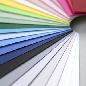 Op de afbeelding: Een waaier van kleurrijke papieren vellen, gerangschikt in een regenboogpatroon, met kleuren die overgaan van rood naar geel, groen, blauw en dan naar wit.