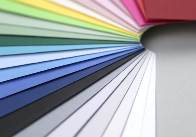 Op de afbeelding: Een waaier van kleurrijke papieren vellen, gerangschikt in een regenboogpatroon, met kleuren die overgaan van rood naar geel, groen, blauw en dan naar wit.