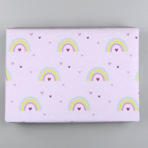 Puede incluir: Papel de regalo rosa con un patrón repetido de arcoíris y pequeños corazones. Los arcoíris son amarillos, azul claro y morados. Los corazones son de un tono morado más oscuro.