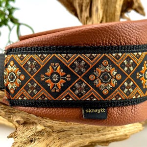 Könnte beinhalten: Ein braunes Leder-Halsband für Hunde mit einem schwarz-orangenen Musterband. Das Band hat ein geometrisches Design mit floralen Akzenten. Das Halsband hat einen silberfarbenen Metallring und ein kleines schwarzes Etikett mit dem Text "skreytt".