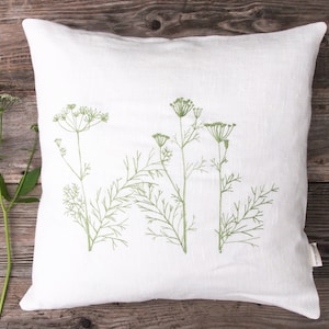 Puede incluir: Almohada de lino blanco con flores silvestres bordadas en verde. La almohada está sobre una superficie de madera.