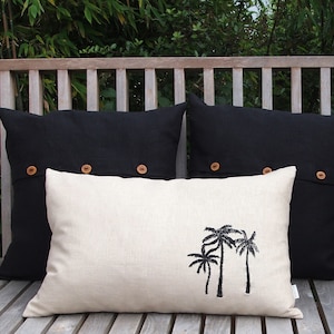 May include: Three decorative throw pillows on a wooden bench. Two black linen pillows with brown buttons and one off-white linen pillow with an embroidered palm tree design.