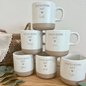 Könnte beinhalten: Sechs weiße Keramikbecher mit braunem, gesprenkeltem Überzug und einem braunen Band um den Boden. Jeder Becher trägt den Text "ERZIEHERIN MIT" und ein Herzsymbol, gefolgt von einem Namen: Isabella, Vanessa, Jofina, Stefanie, Amelie und Bettina.