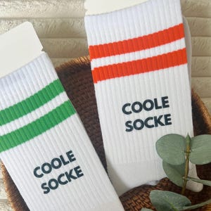 Könnte beinhalten: Zwei Paar weiße Socken mit farbigen Streifen und dem Text "COOLE SOCKE". Ein Paar hat grüne Streifen, das andere orangefarbene Streifen. Die Socken werden in einem Weidenkorb mit Grünzeug präsentiert.