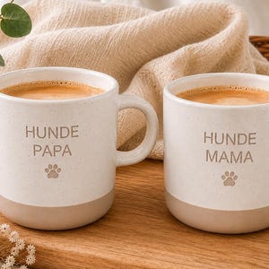 Könnte beinhalten: Zwei cremeweiße Keramiktassen mit gesprenkelter Oberfläche und hellbraunem Boden. Jede Tasse hat einen Henkel und ist mit Kaffee gefüllt. Die linke Tasse trägt den Text HUNDE PAPA mit Pfotenabdruck, die rechte Tasse HUNDE MAMA mit Pfotenabdruck.