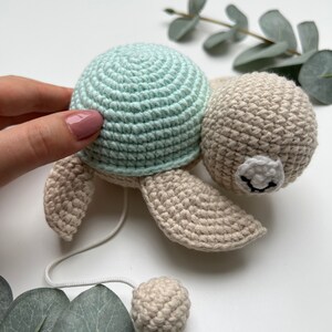 Pu&ograve; includere: Un giocattolo a forma di tartaruga all'uncinetto con un guscio verde menta chiaro e un corpo beige. La tartaruga ha una testa rotonda con una macchia bianca e tratti del viso ricamati in nero. Un cordino bianco parte dal corpo verso una piccola sfera beige.