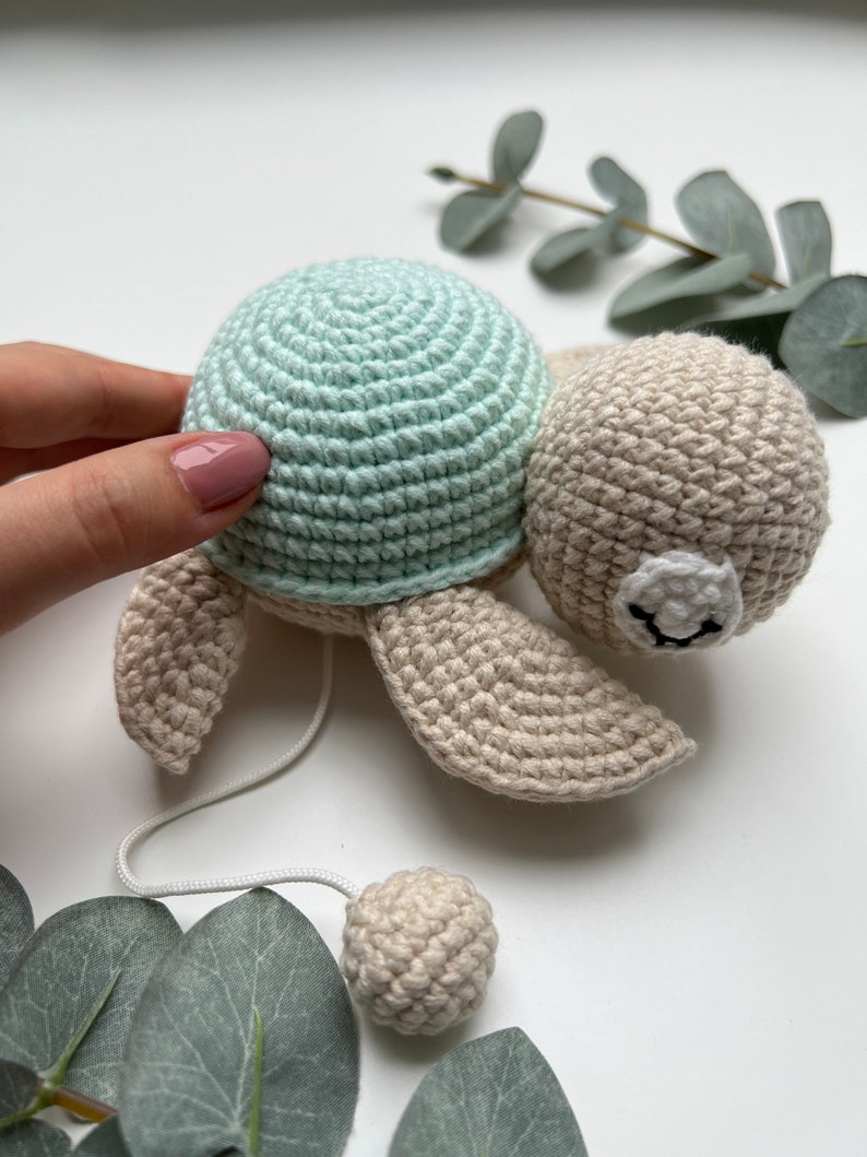 Pu&ograve; includere: Un giocattolo a forma di tartaruga all'uncinetto con un guscio verde menta chiaro e un corpo beige. La tartaruga ha una testa rotonda con una macchia bianca e tratti del viso ricamati in nero. Un cordino bianco parte dal corpo verso una piccola sfera beige.