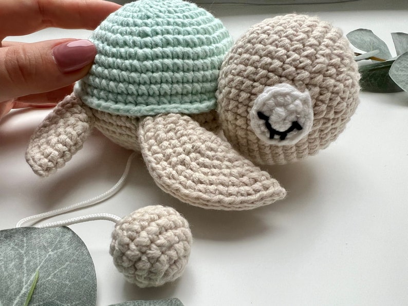 Pu&ograve; includere: Un giocattolo a forma di tartaruga marina all'uncinetto con un guscio verde menta chiaro e un corpo beige. La tartaruga ha una testa rotonda con una toppa circolare bianca sul viso, ricamata con una bocca sorridente nera. Una piccola pallina all'uncinetto beige pende da un cordino bianco.