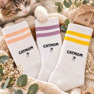 Könnte beinhalten: Drei Paar weiße Socken mit gerippten Bündchen und zwei horizontalen Streifen in Pfirsich, Lavendel und Gelb. Jede Socke zeigt den schwarzen Schriftzug CATMOM über einem kleinen schwarzen Pfotenabdruck-Symbol mit zwei vertikalen Linien.