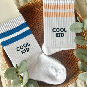 Op de afbeelding: Twee paar witte sokken met de tekst "COOL KID". Eén paar heeft blauwe strepen, de andere heeft perzikstrepen. De sokken worden gepresenteerd in een rieten mand met groen.