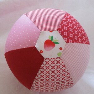 Könnte beinhalten: Ein roter, rosa und weißer Patchwork-Ball mit einem Erdbeer-Design. Der Ball ist aus Stoff und hat eine weiche, plüschige Textur.