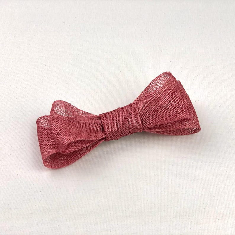 May include: A burgundy-colored bow made of a woven material. The bow is tied in the center and has two loops on each side. The bow is lying on a white surface.