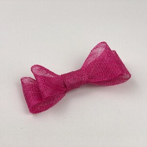 May include: A bright pink bow made of a mesh-like fabric. The bow is tied in the center and has two loops on each side. The bow is laying on a white surface.