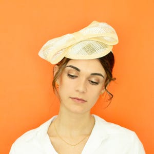 May include: A woman wearing a white shirt and a cream-coloured straw hat with a bow. The hat is tilted slightly to the side. The woman is looking down.